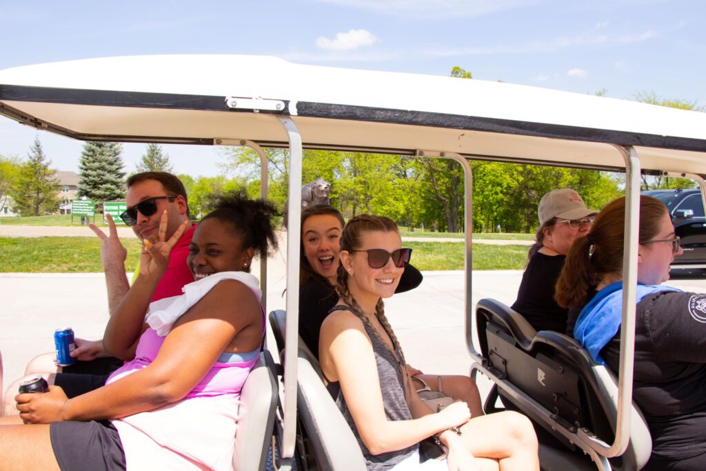 A group of six people, three of them visibly joyful, riding in a golf cart on a sunny day, with green trees in the background.