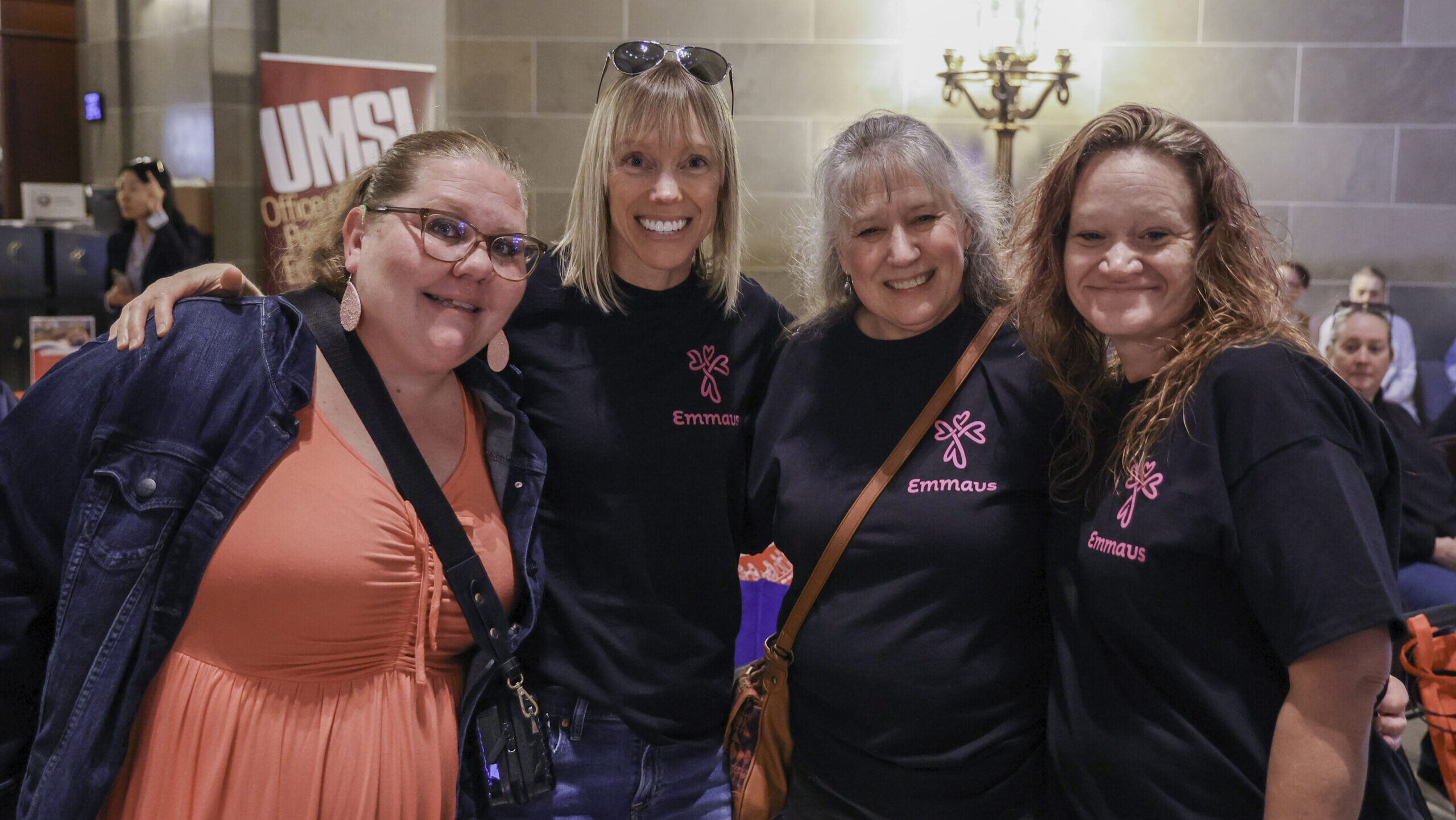 Four women smiling and posing together at an event. They are wearing Emmaus t-shirts, and one of them is also wearing a coral-colored dress and glasses. The background shows a UMSL banner and a well-lit interior.