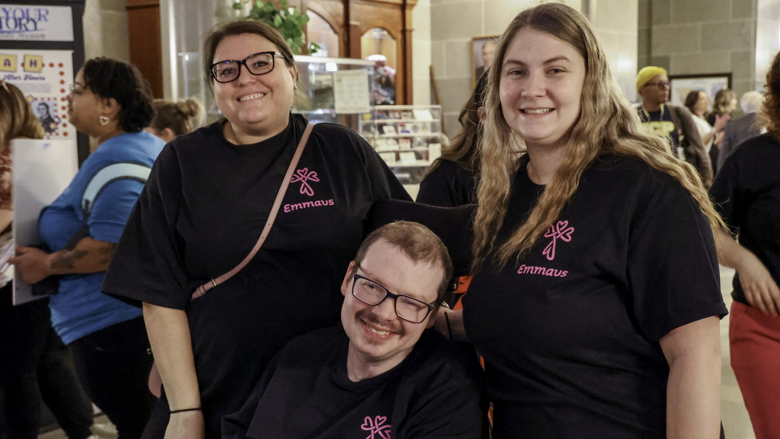 Two DSPs and on clients in black Emmaus t-shirts smile for the camera; one is seated in a wheelchair, at MO Capital for DRLD Day.