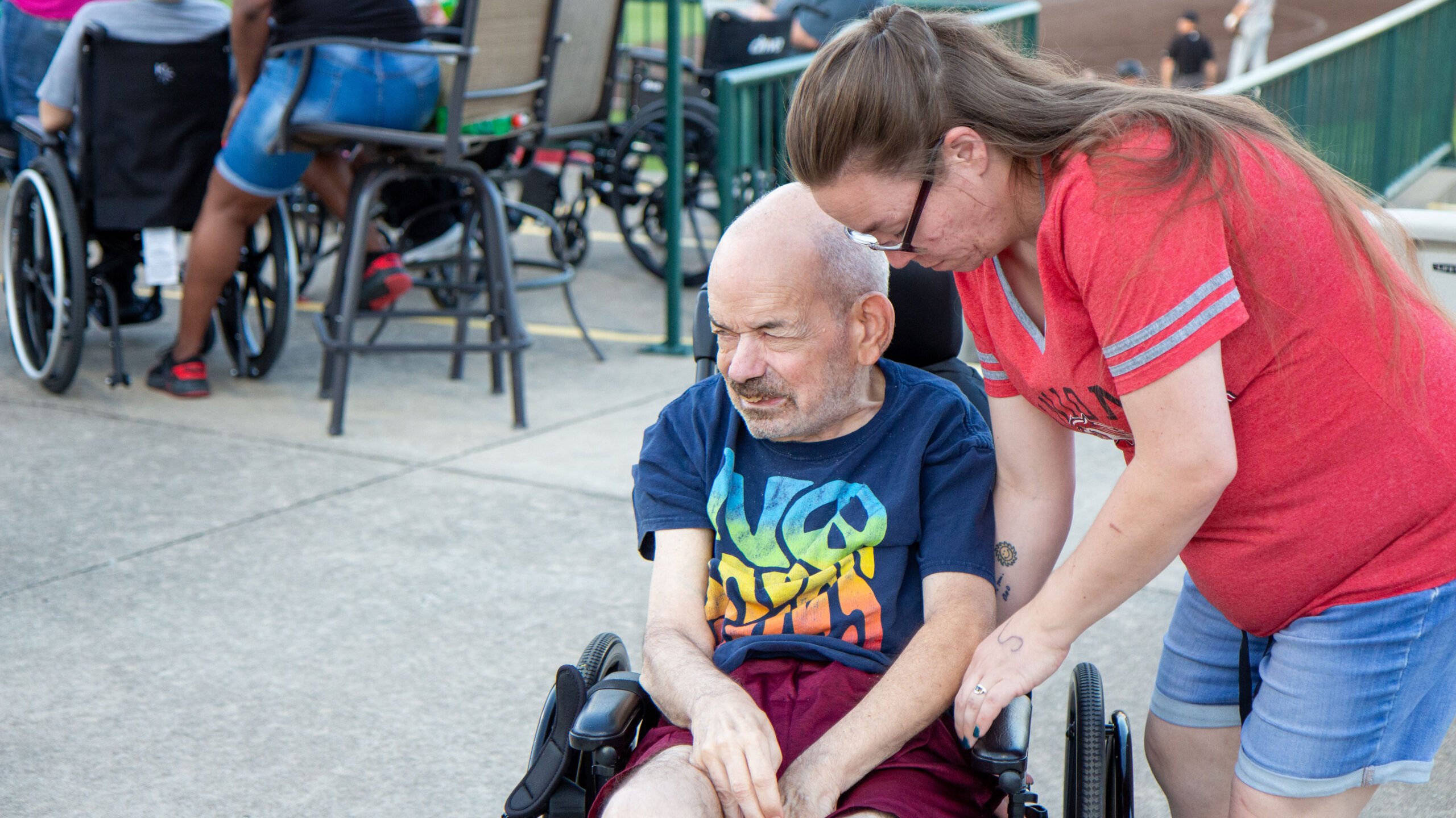  A woman in a red shirt leaning over to talk to an older man in a wheelchair. The man is wearing a colorful t-shirt and red shorts. Other people in wheelchairs and chairs are visible in the background.