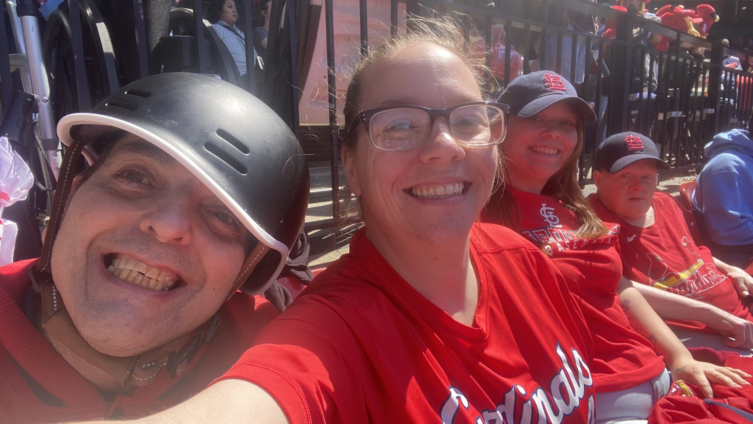 Four Emmaus Clients and DSPs in red Cardinals attire smile and enjoy a sunny day at a baseball game; one is wearing a helmet