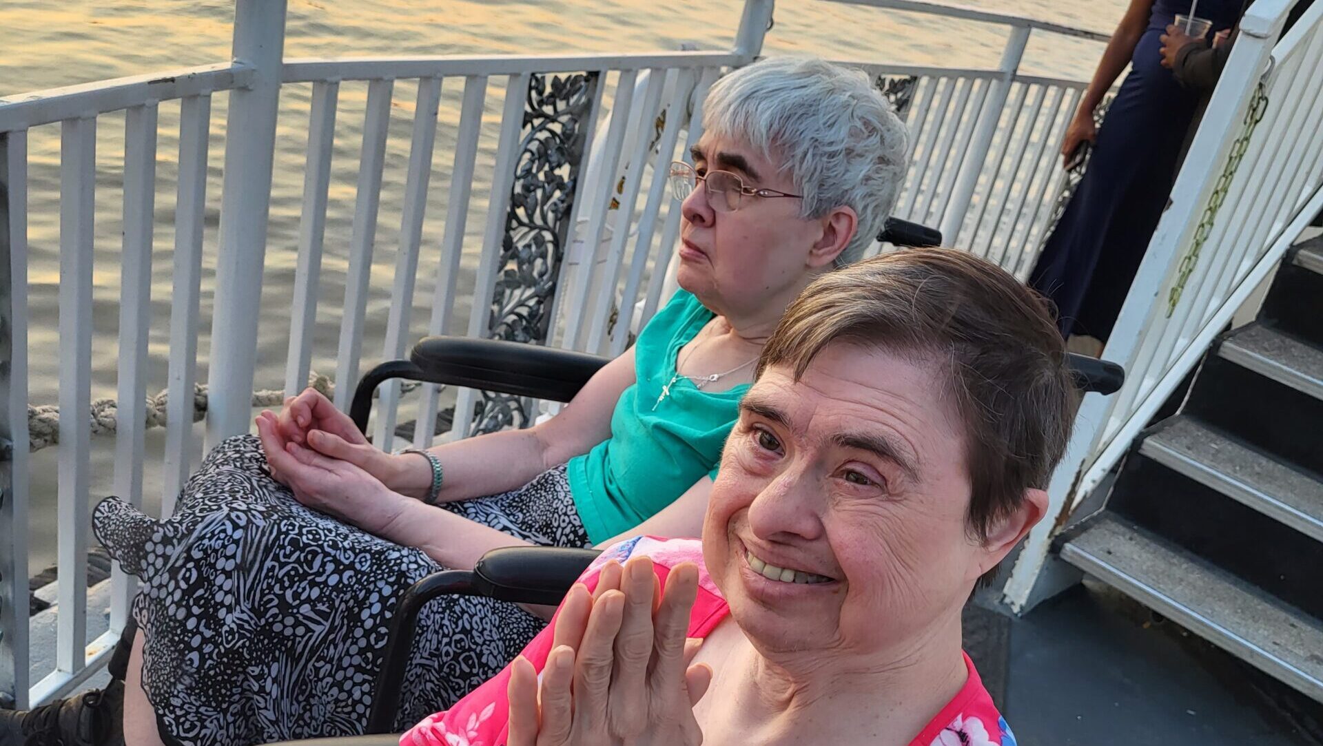 Two women in wheelchairs, one in a floral dress and the other in a turquoise top, enjoy a peaceful river view from a boat deck.