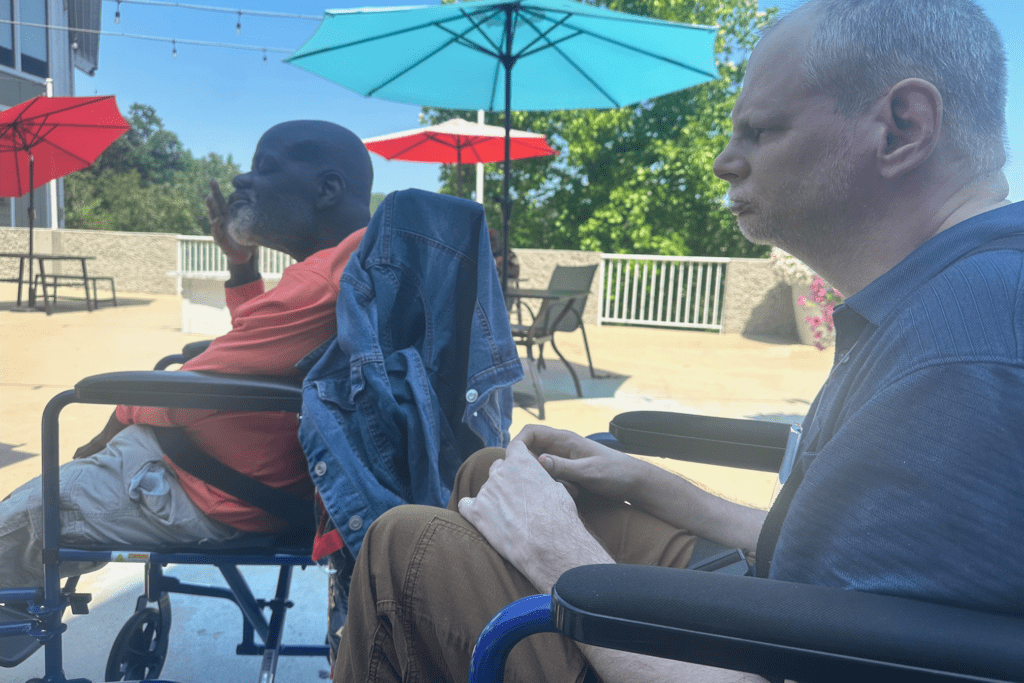 Two men sit in wheelchairs outside on a sunny patio with colorful umbrellas overhead. One man wears an orange shirt, the other a blue shirt, with a denim jacket draped over his chair.