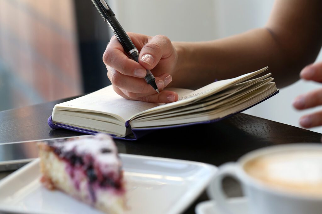  Close-up of a person’s hand writing in a journal with a black pen. The journal is open and resting on a table in a café, with a piece of berry-topped cheesecake on a white plate and a cup of coffee in the foreground