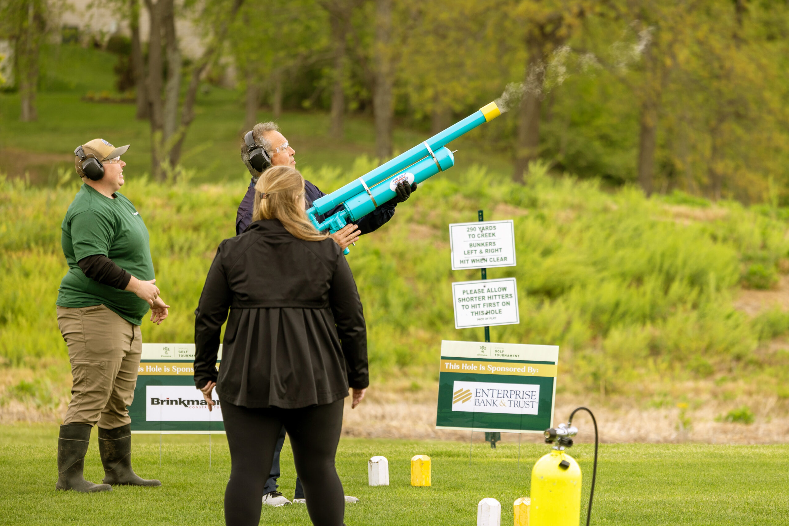 A golfer at the Emmaus golf tournament aims and fires a bright blue golf ball launcher while wearing protective ear gear. Two other participants, also wearing ear protection, watch nearby on the green