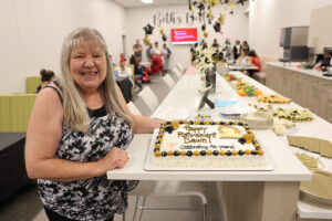 A retirement cake for Dawn, decorated with gold and black icing, featuring her photo and the message, "Happy Retirement Dawn! Celebrating 46 years!" The cake sits on a table with plates, napkins, and a spread of party food.