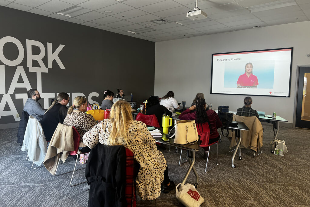 A group of new Direct Support Professionals (DSPs) attending a training session at The Learning Lab at Emmaus. They are seated at tables watching a training video on recognizing choking, projected on a large screen. The room has a modern design with "WORK THAT MATTERS" displayed on the wall.