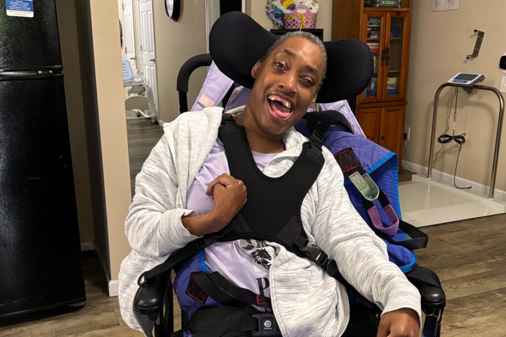 Smiling woman sitting upright in a supportive wheelchair, wearing a gray hoodie, in a bright and welcoming living space.