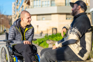 A man in a wheelchair smiling and chatting with a friend on a park bench.