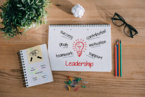 Top view of a desk with an open notebook labeled “Leadership” and related words, plus a smaller notepad, plant, glasses, and stationery.